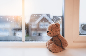 Lonely bear toy sitting alone looking out of window, Sad teddy bear doll sitting next to window in...