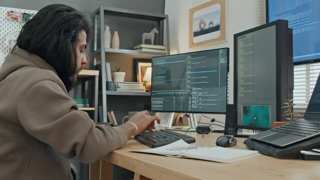 Medium fast motion shot of workday of busy young male software developer working from home - typing computer code on large screen, writing in notebook and drinking coffee