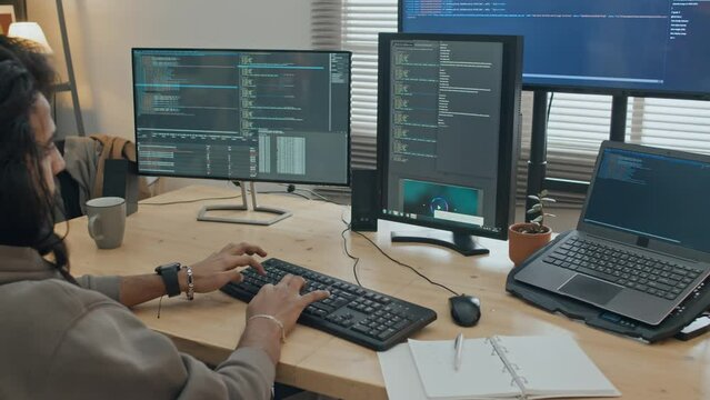 Medium arc shot of young man with long hair and beard, in sweatshirt sitting at desk at home or in office, and typing on keyboard, while writing computer program in coding software on large screen