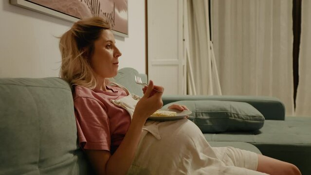 Side view of female with an appetite holds plate and eating dessert while pregnant on sofa. Irresistible craving during late pregnancy a woman on the couch binging cake.