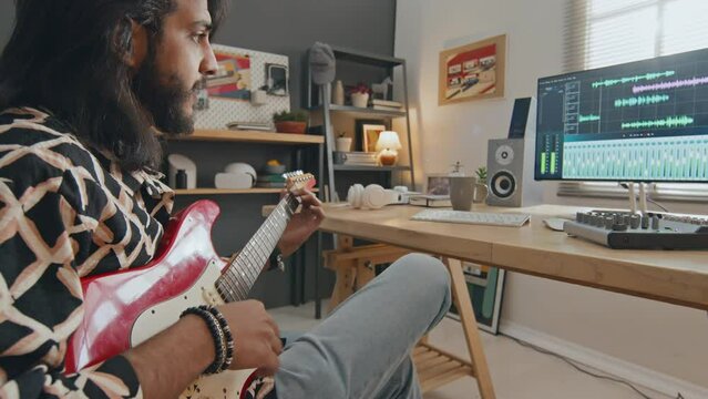Medium zoom shot of young male Middle Eastern musician with beard and long hair sitting at desk with computer, playing song on electric guitar and recording music with audio editing software