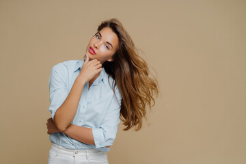 Reflective young woman in a blue shirt, looking upwards with flowing hair, against a beige...