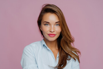 Fototapeta premium Stunning woman with flowing hair and blue shirt, offering a soft smile against a pink background