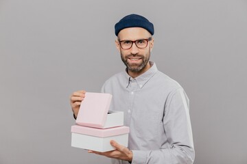 Handsome European man with thick stubble, pleasant smile, holds boxes in hands, opens gift with happiness, wears formal white shirt and headgear, isolated over grey background. Parcel in mans hands