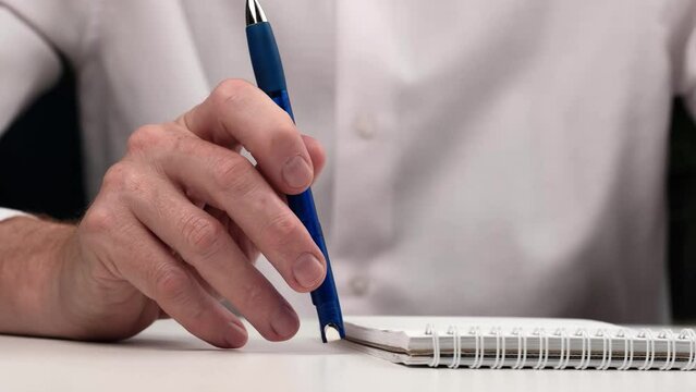 man slowly and thoughtfully taps a blue pen on table next to notebook. closeup.