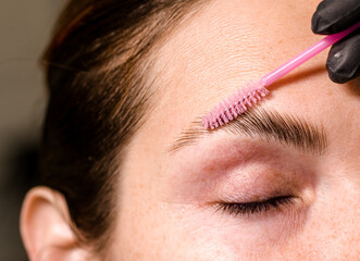 Eyebrow tinting, a young woman sitting in a chair at a brow artist's station. The artist is...
