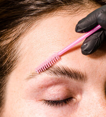 Eyebrow tinting, a young woman sitting in a chair at a brow artist's station. The artist is applying dye to the woman's eyebrows.