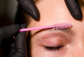 Eyebrow tinting, a young woman sitting in a chair at a brow artist's station. The artist is...