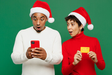 Merry young couple friend man woman wear red casual clothes Santa hat posing using mobile cell phone credit bank card isolated on plain green background. Happy New Year 2024 Christmas holiday concept.