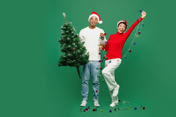 Full body surprised fun merry young couple man woman wear red casual clothes Santa hat posing hold Christmas tree with garlands isolated on plain green background. Happy New Year 2024 holiday concept.