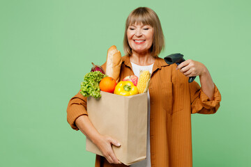 Elderly smiling happy satisfied woman wear brown shirt casual clothes hold shopping paper bag with scanning food products check bar code isolated on plain green background. Delivery service from shop.