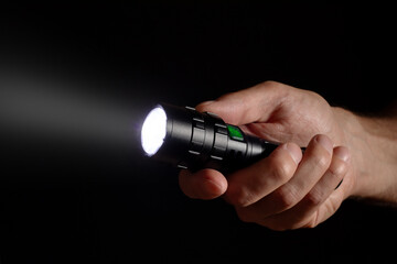 Male hand holding a led flashlight with a wide white beam on a black background, leaving the right...
