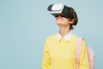 Young smiling happy cheerful woman student wear casual clothes yellow sweater backpack bag watching in vr headset pc gadget isolated on plain blue background. High school university college concept.