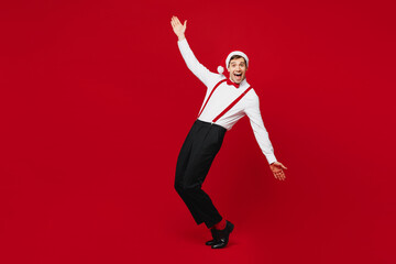 Full body merry young man wear white shirt Santa hat stand on toes leaning back with outstretched hands posing isolated on plain red background. Happy New Year Christmas celebration holiday concept.