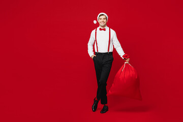 Full body merry young fun man wear white shirt Santa hat posing hold bag with present gifts look...