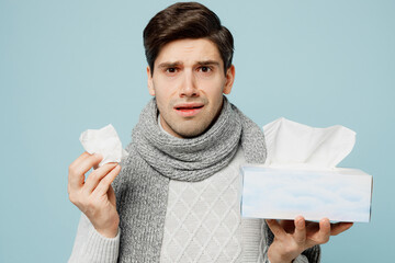 Young ill sick man wear gray sweater scarf sneezing hold box of paper napkins isolated on plain...