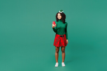 Full body merry little kid teen girl wear hat casual clothes posing hold in hand use mobile cell phone isolated on plain green background studio. Happy New Year celebration Christmas holiday concept.