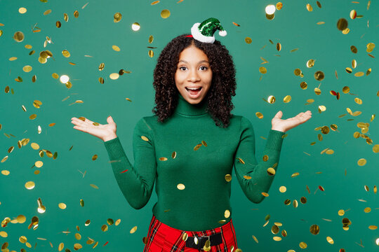Merry fun excited little kid teen girl wear hat casual clothes posing spread hands in confetti rainfall isolated on plain green background studio. Happy New Year celebration Christmas holiday concept.