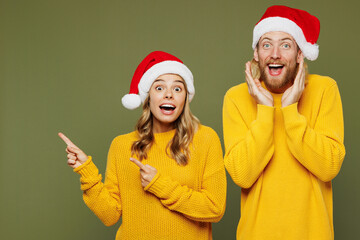 Merry surprised young couple two friends man woman wear sweater Santa hat posing point index finger aside on area hold face isolated on plain green background Happy New Year Christmas holiday concept