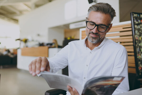 Male happy calm rich smiling confident businessman man wears classic shirt read catalog sitting at office table in dealership store indoors want to buy new automobile car. Business lifestyle concept.