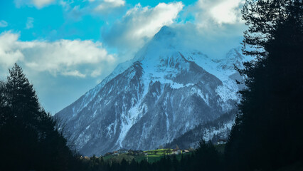 A massive mountain peak rising above the shaded forests. The snowed crest touches the clouds. Swiss Alps.  - obrazy, fototapety, plakaty