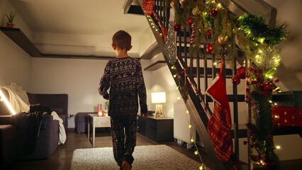 Little boy in pajamas at night walking down the wooden stairs at house decorated for Christmas or New Year celebrations