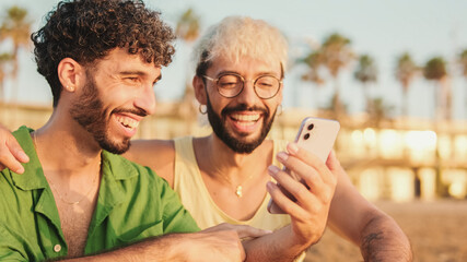 Close-up of young gay couple sitting embracing, smiling and laughing have fun with mobile phone...
