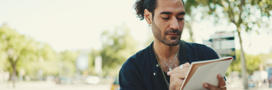 Fototapeta Young italian guy with ponytail and stubble sits on street bench and makes sketches with pen on piece of paper