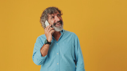Elderly grey-haired bearded man wears a blue shirt, annoyed and frustrated talking on a mobile...