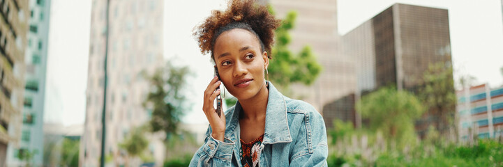 Beautiful African girl with ponytail wearing denim jacket talking on mobile phone on modern city...