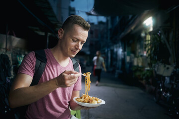 Tourist at night Bangkok. Man with backpack eating local food (pad thai) at street market..