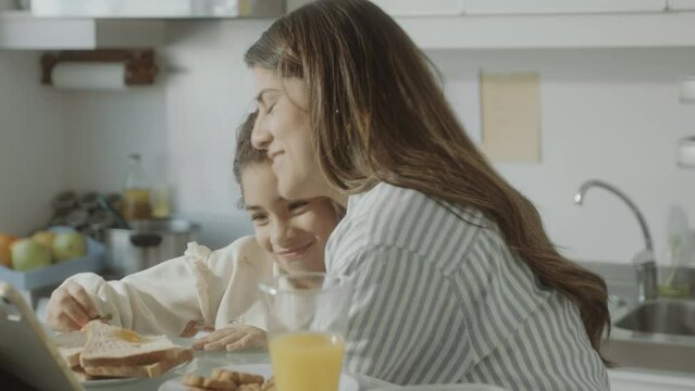 Loving mother talking with adorable little daughter over breakfast at home, then hugging and kissing her
