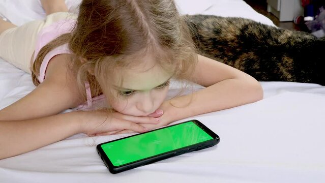 A child looks at a green phone screen in bed with a black cat. Chromed green screen. Communication, game or cartoon on the phone