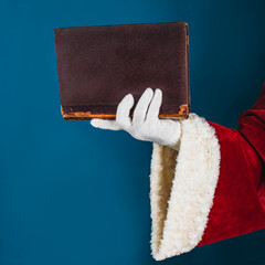 Santa's hand in a white glove holds a old book on a turquoise background with copy space. Concept for knowledge, education, history, faith, literature, read, leisure.
