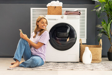 Beautiful smiling woman, housewife holding mobile phone sitting on carpet looking away in modern laundry room with washing machine, laundry detergent, basket with clothes on background. Advertisement - Powered by Adobe