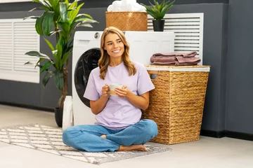 Fotobehang Smiling woman, housekeeper holding cup of coffee sitting in lotus position at cozy home near washing machine. Cleaning, laundry concept © Maria Vitkovska