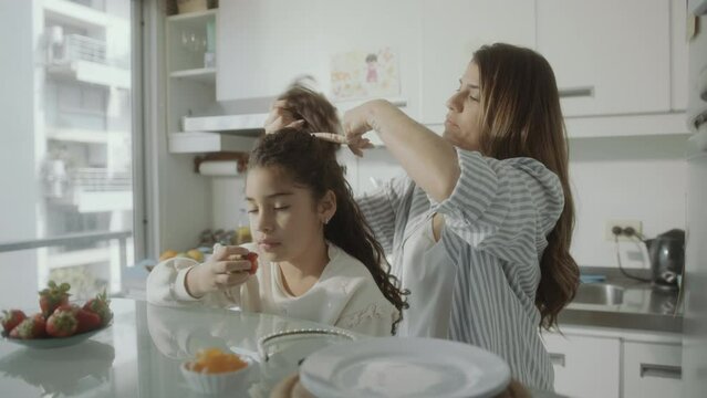 Young careful mother tying curly hair of little daughter in a ponytail as girl eating strawberry in the kitchen at home