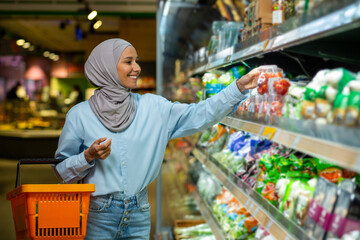 Joyful smiling Arab woman in a hijab chooses products in a supermarket, walks with a basket in her...