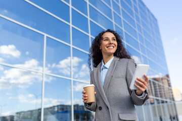 Successful Hispanic businesswoman walks to the office, holds a coffee and a phone in her hands,...