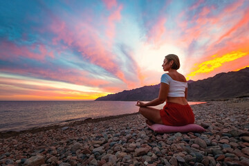 Young woman shilhouette in meditation position on the beach against sunset sky