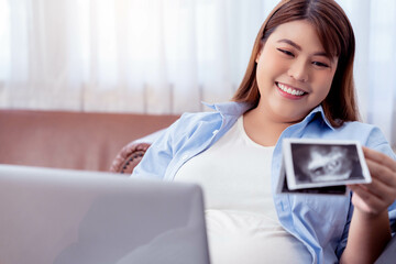 Asian pregnant woman working on a laptop look at ultrasound picture of her baby Young beautiful...