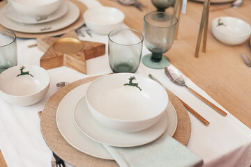 minimal scandinavian trendy Christmas table setting of white plates with green reindeer print, napkin and wooden table and decor cutlery