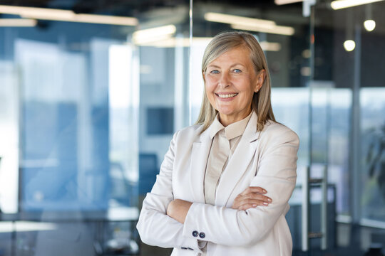 Portrait of mature successful senior woman boss inside office in business suit, businesswoman smiling and looking at camera, standing by window with crossed arms, banker finance woman.