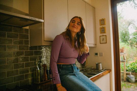 Young woman in fashionable outfit sitting on kitchen counter and smiling