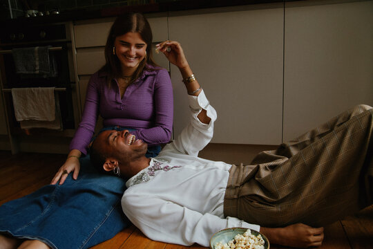 Young non-binary person and woman having fun hanging out on kitchen floor eating popcorn