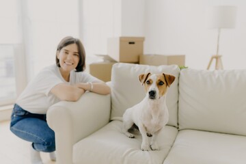 Smiling woman and her dog on a white sofa with moving boxes in a light-filled living room