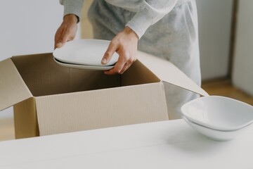 Hands carefully unpacking white dishes from a cardboard box on a table in a bright room.