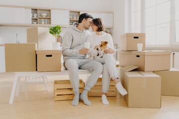 Affectionate couple with a dog enjoying a coffee break amid moving boxes in a bright kitchen