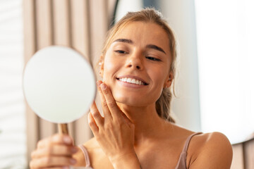 Closeup of attractive smiling woman touching her cheek applying face cream looking in mirror....