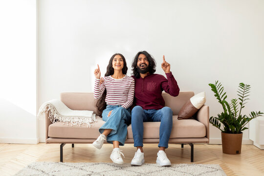 Excited eastern spouses sitting on couch, pointing up